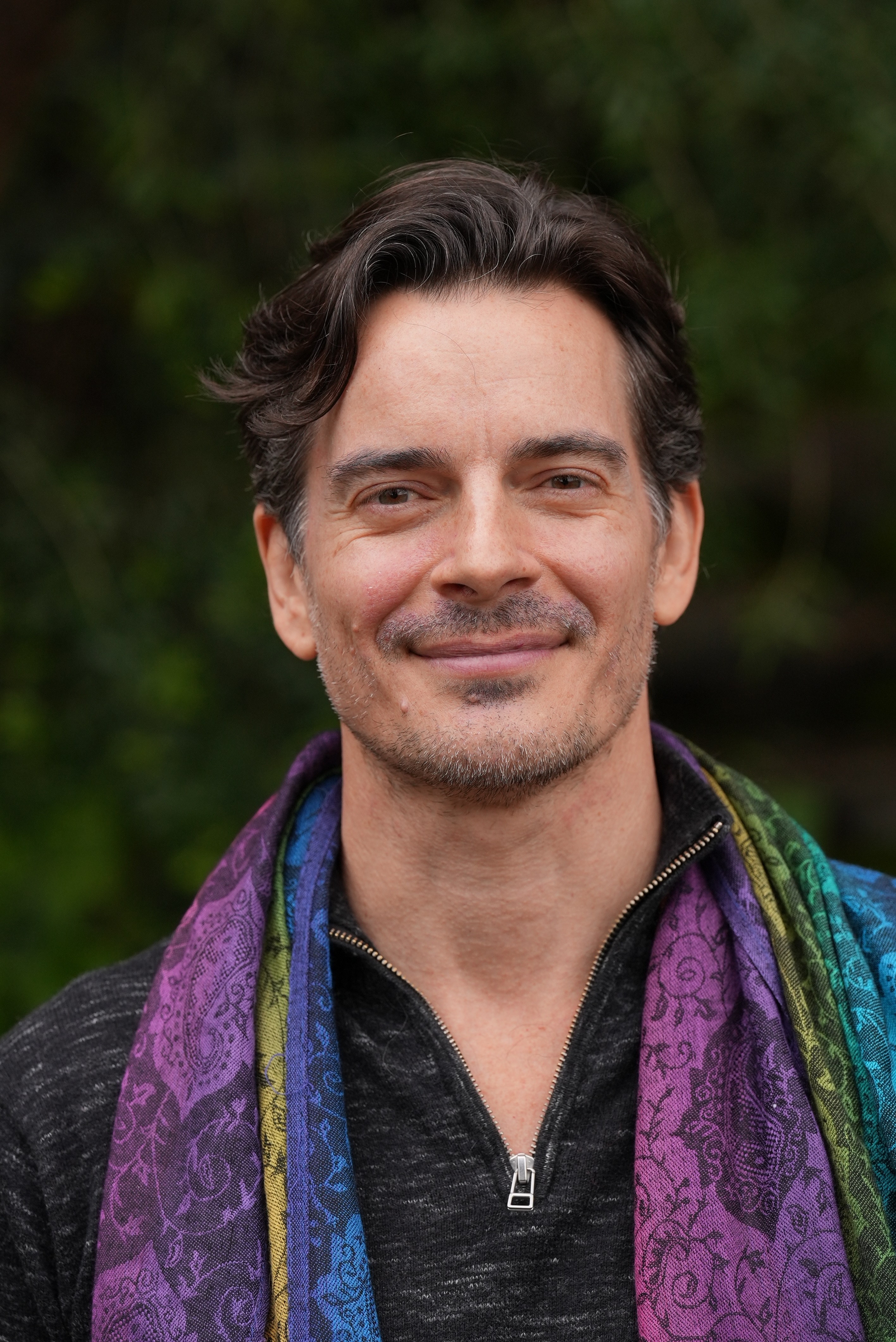 Portrait of Romie Kind, principal of The Life Robotic: head and shoulders, outdoors against green foliage, dark-grey quarter-zip with a paisley scarf in purple, teal, and chartreuse, warm direct expression.