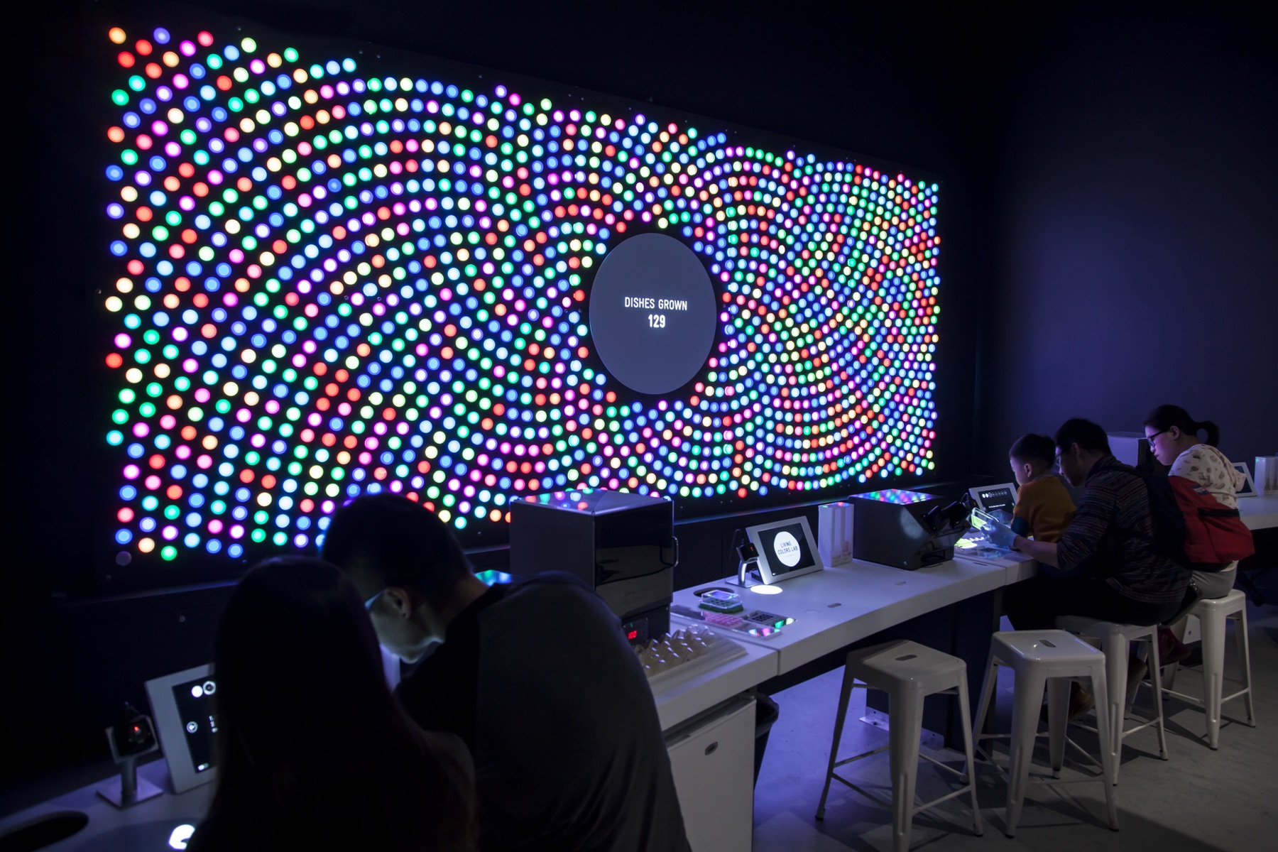 The Living Colors Lab pigment wall: a large grid of red, green, and blue lights forming spiral patterns, with a central counter reading the number of bacterial dishes grown. Visitors work at lit benches below it.