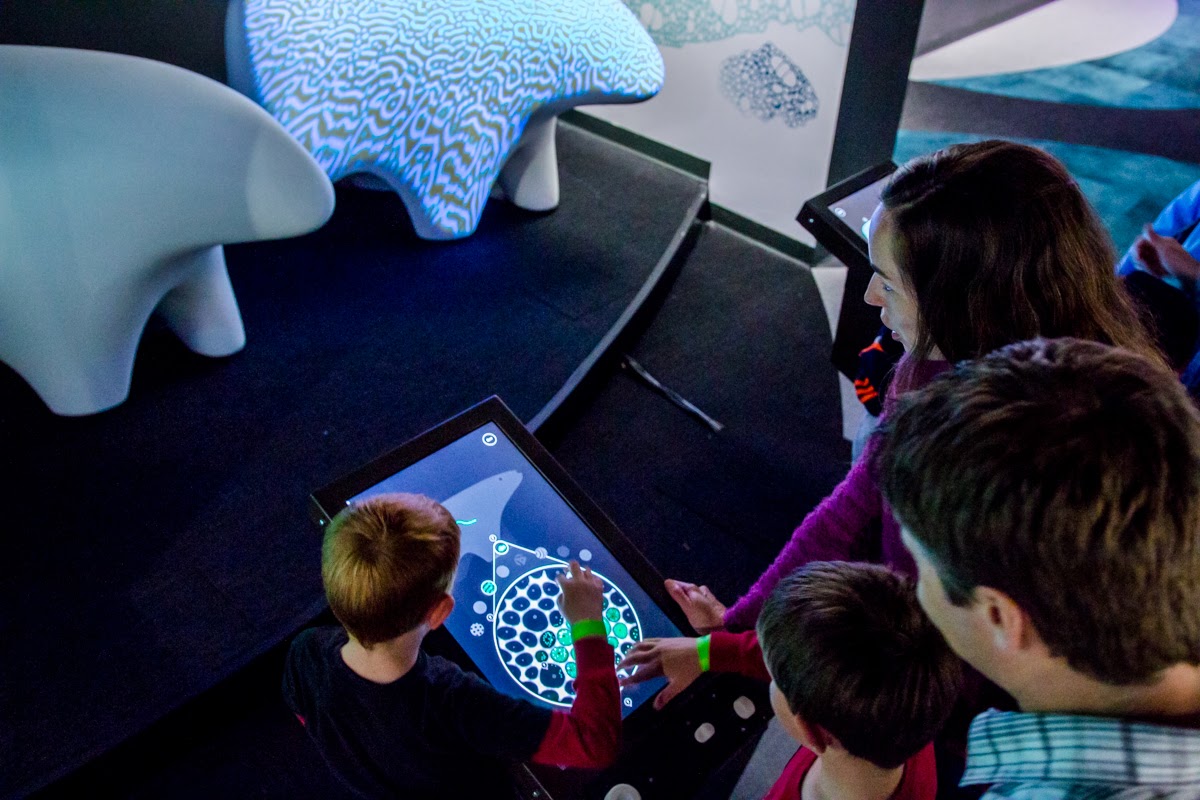 Three young visitors and an adult lean over an interactive tabletop in BioDesign Studio. The screen shows a circular cell-design interface. In the background, an organic-form sculpture printed with a generative pattern is partly visible.