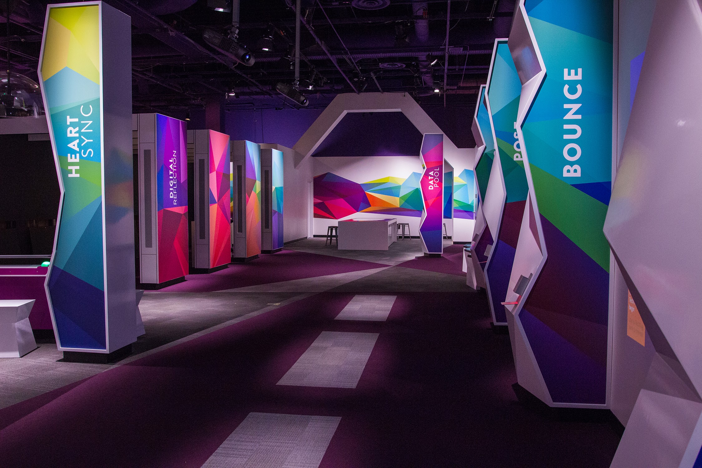 The Body Metrics exhibition floor at The Tech Interactive: a procession of tall faceted columns in cyan, magenta, blue, and teal labeled HEART SYNC, DIGITAL REFLECTION, DATA POOL, and BOUNCE, set against a deep purple carpet under museum lighting.