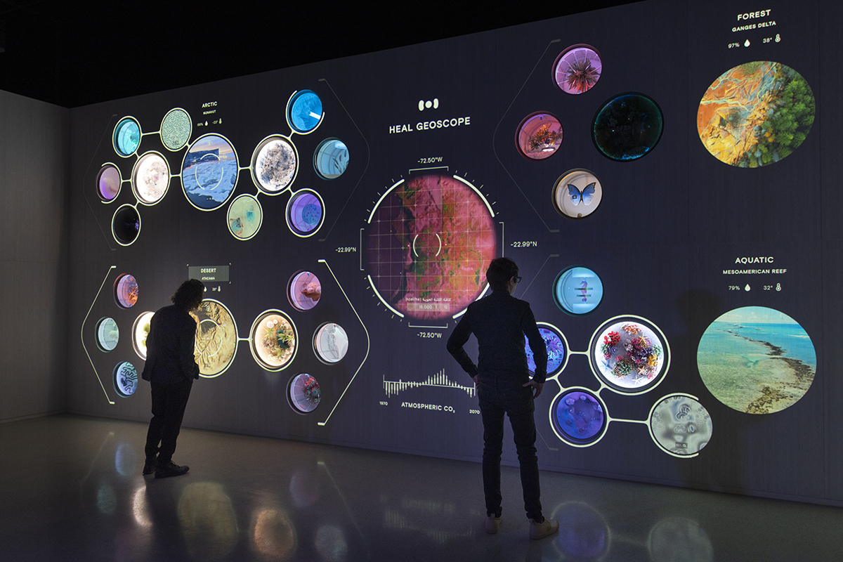 Two figures silhouetted against the HEAL Geoscope wall at the HEAL Institute pavilion. The wall shows a hexagonal grid of circular ecology displays for Arctic, Forest, and Aquatic zones, with a central radial visualization of atmospheric CO2.